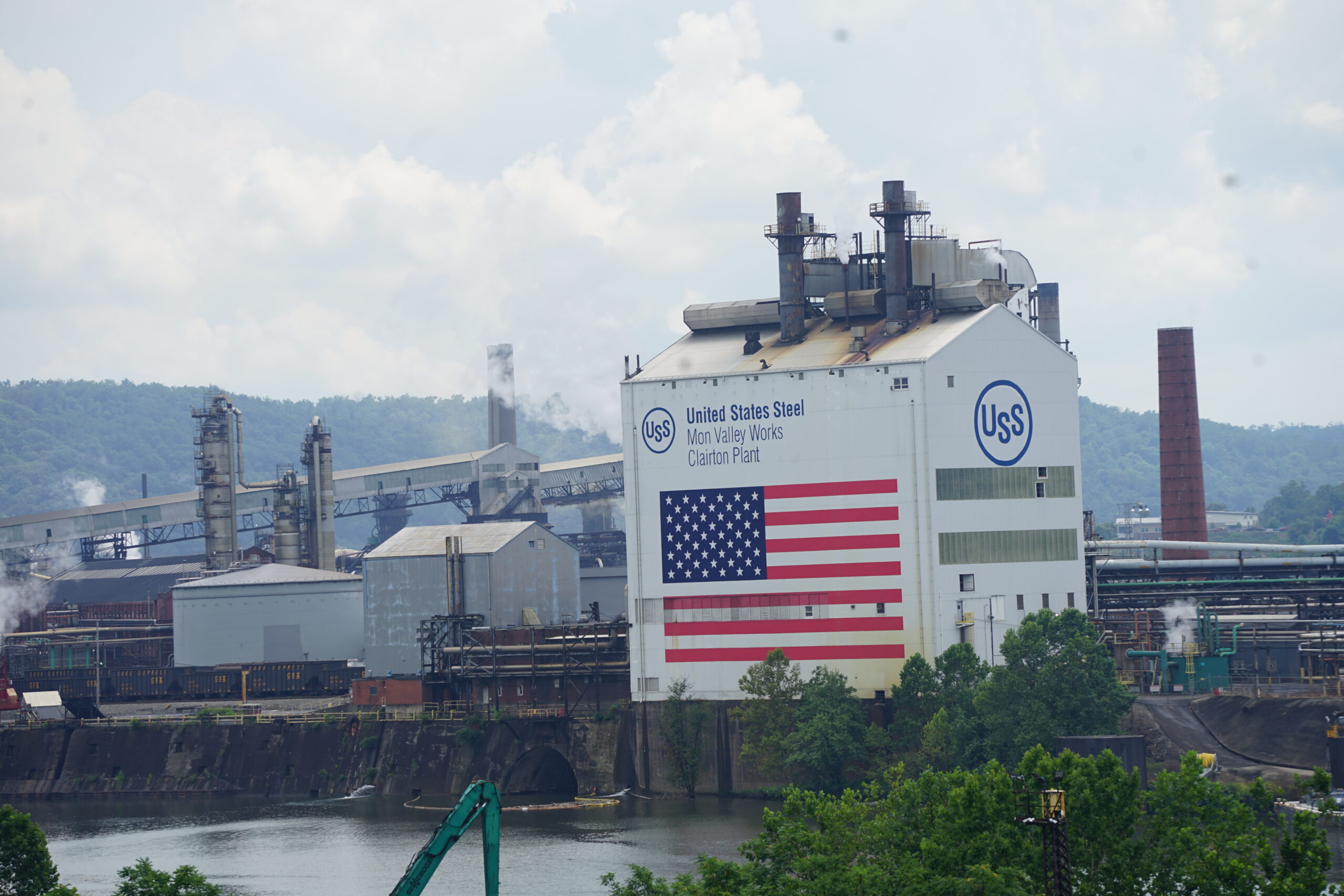 A sprawling industrial facility with a large American flag on the side of a building, along a river.