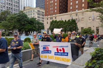 People walk on a street carrying a banner that reads "Safe Jobs Now," with city buildings behind them.