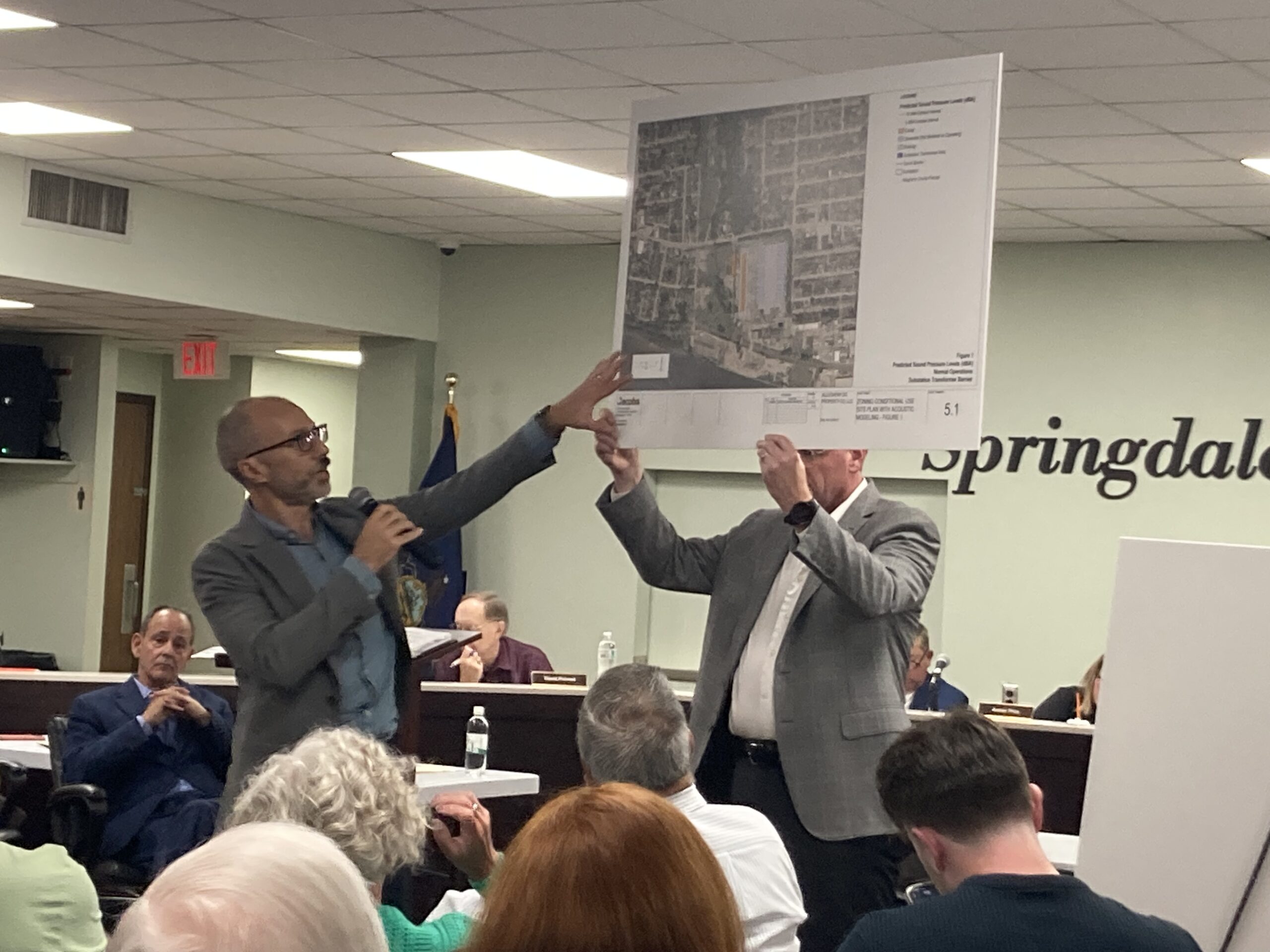 A man points to an arial photo being held up by another man, in front of people seated in chairs