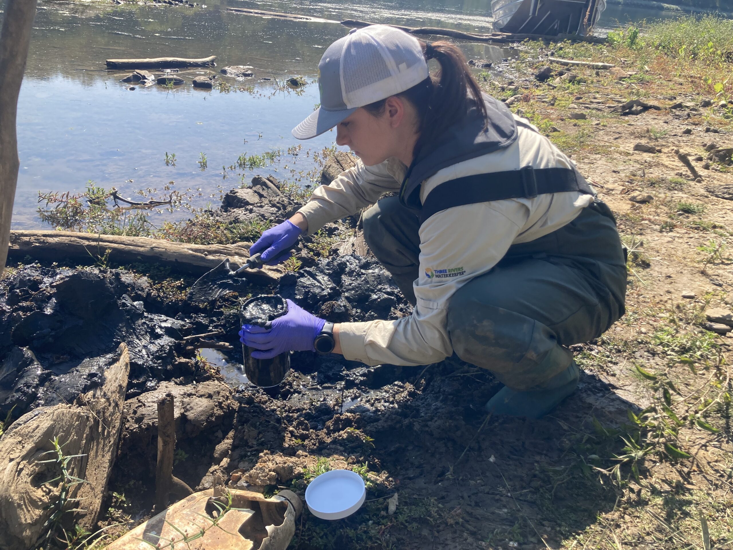 A woman crouching by the river's edge, scooping mud into a glass jar