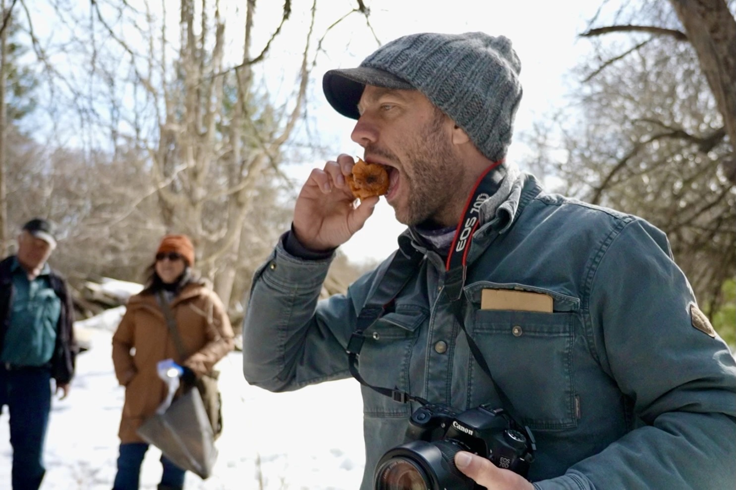 A man takes a bite of an apple in an orchard in the winter as two people look on