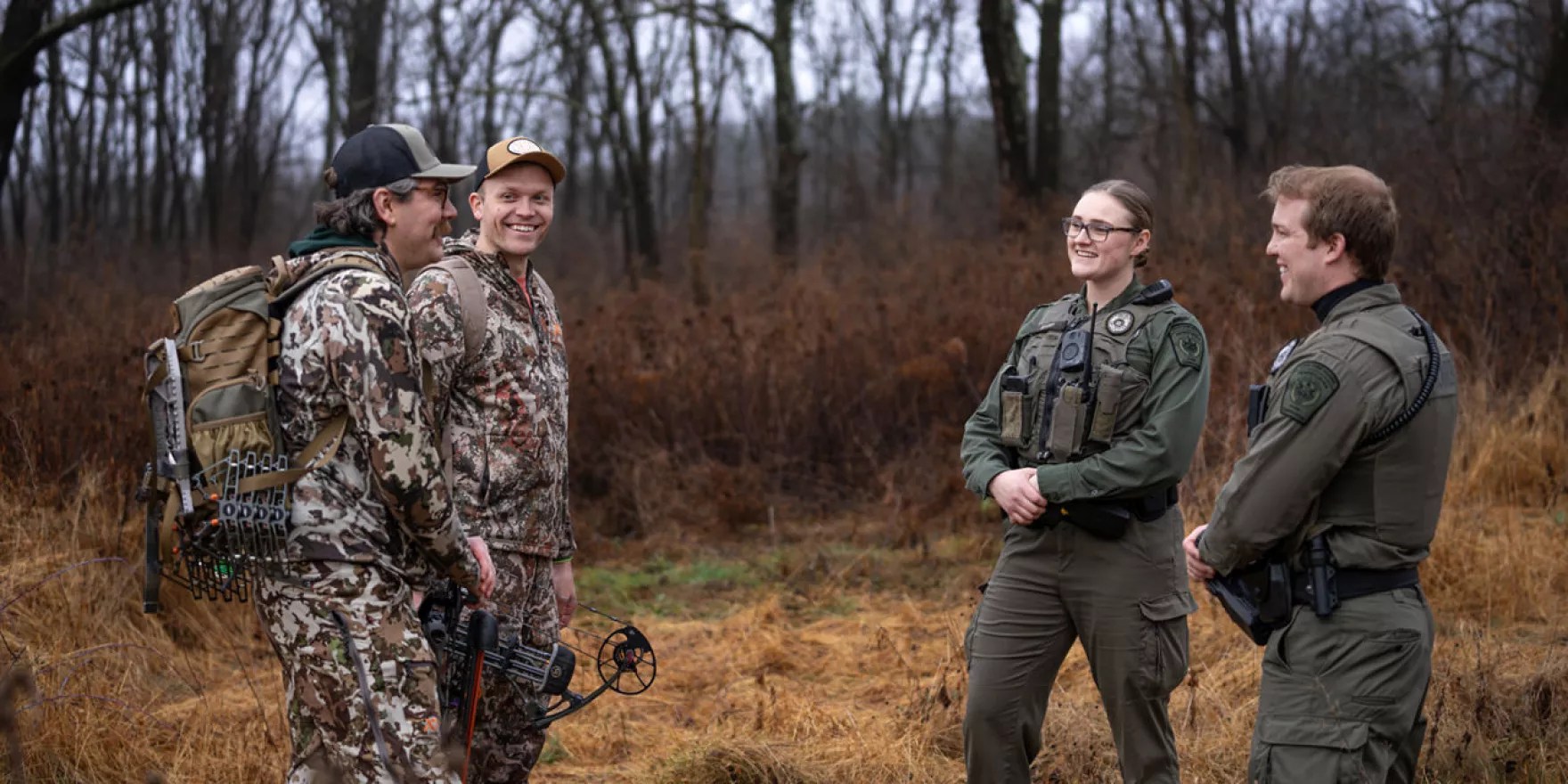In the woods, two wardens stand on the right, talking to two people wearing camouflage.