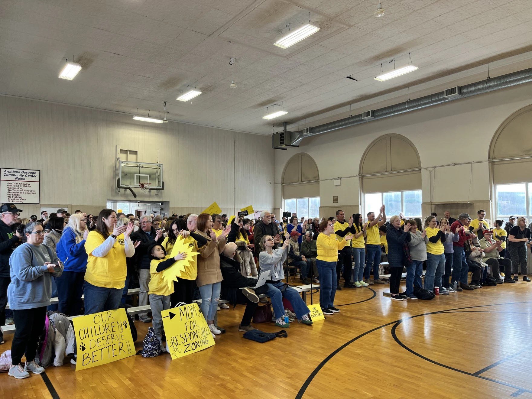 A group of people, some wearing yellow t-shirts with home-made signs, are standing up cheering in a meeting room.