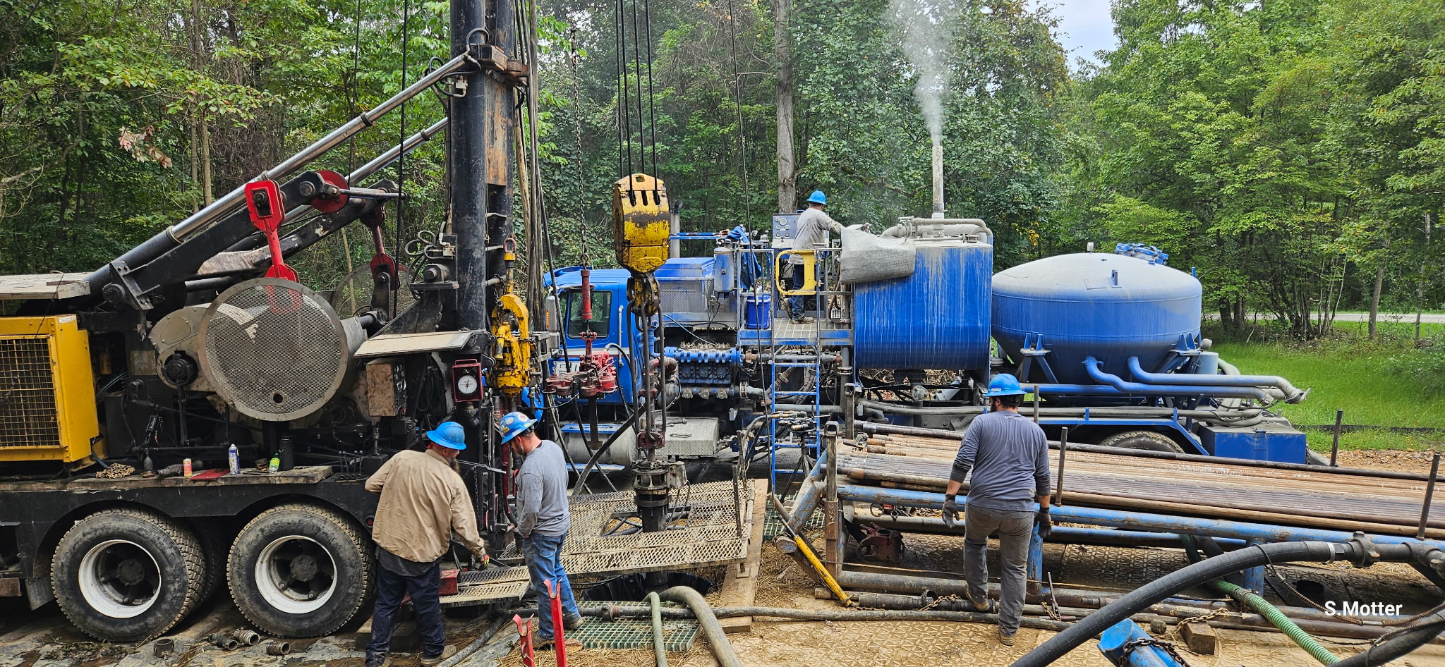 Blue colored industrial eqiupment and workers at a gas well with green trees in the background