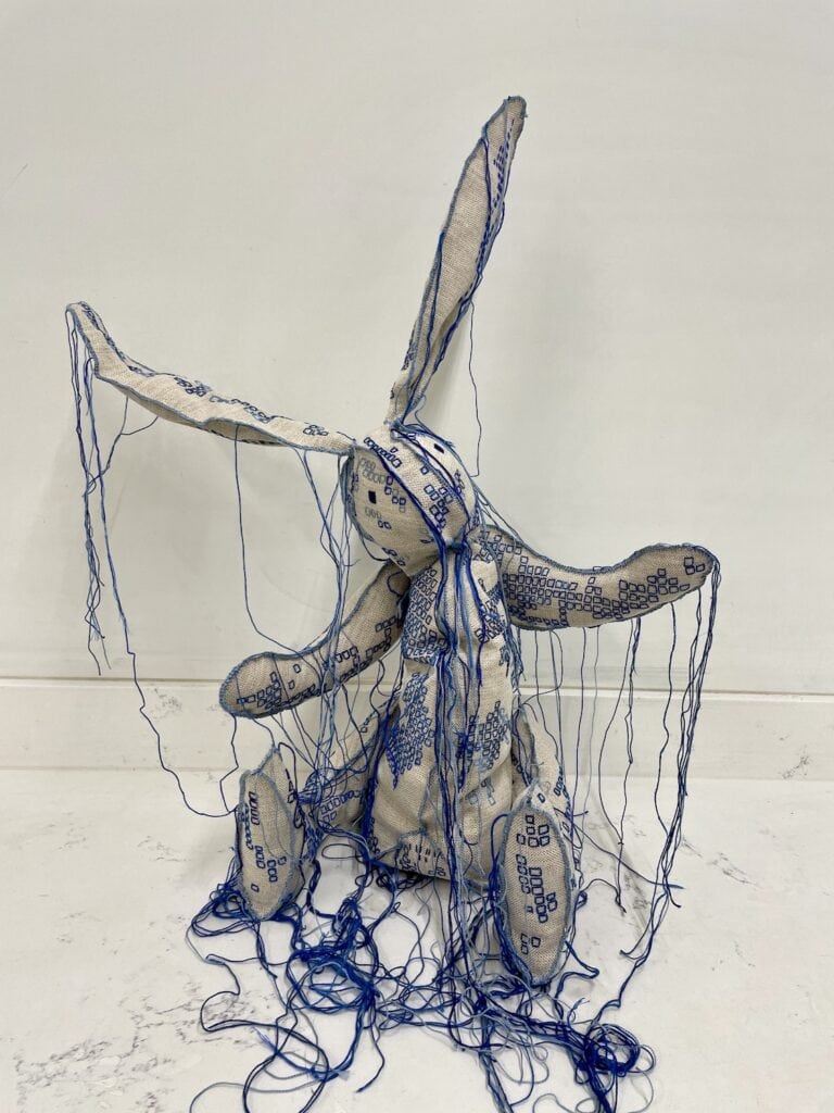 Rabbit 'stuffed animal' - embroidered with squares of different hues of blue, many strings hanging down from the fabric. Sitting on a white marble surface.