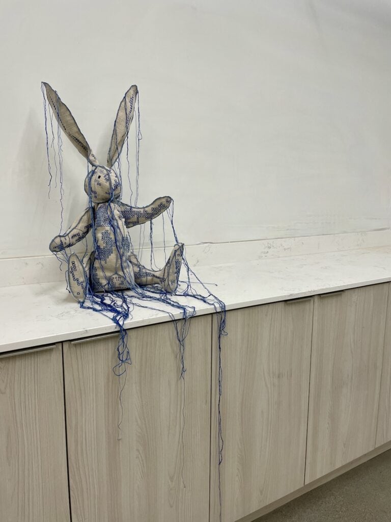 Rabbit 'stuffed animal' - embroidered with squares of different hues of blue, many strings hanging down from the fabric. Sitting on a white marble counter top.