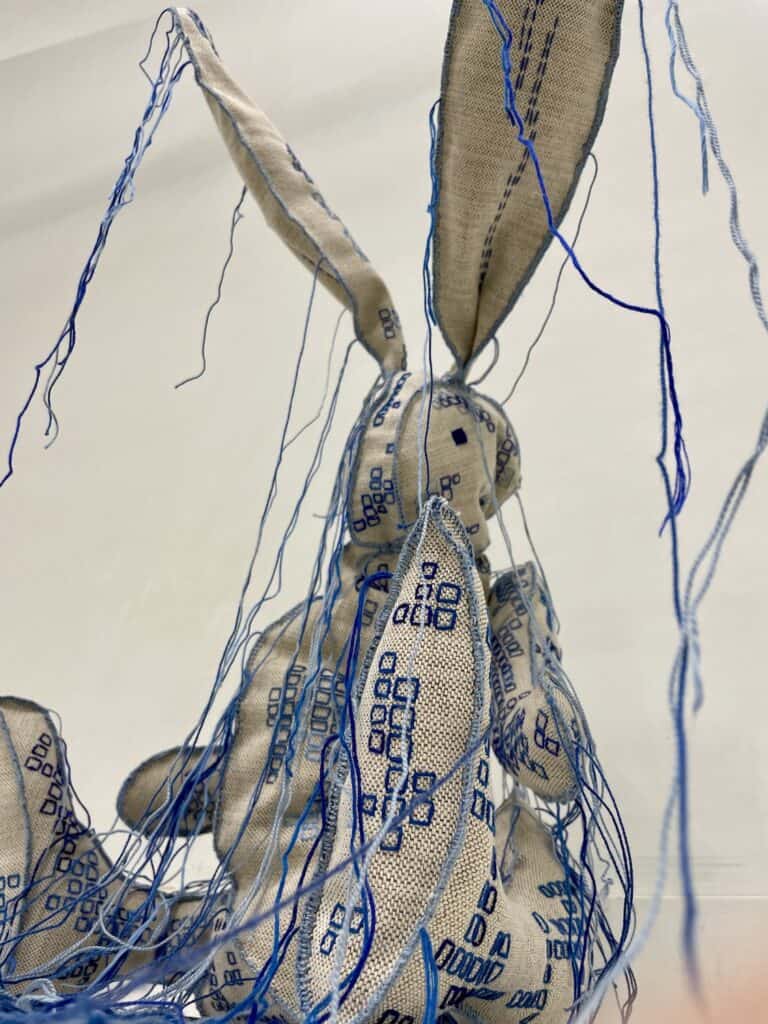 Detail of the food of a Rabbit 'stuffed animal' - embroidered with squares of different hues of blue, many strings hanging down from the fabric. Sitting on a white marble surface.