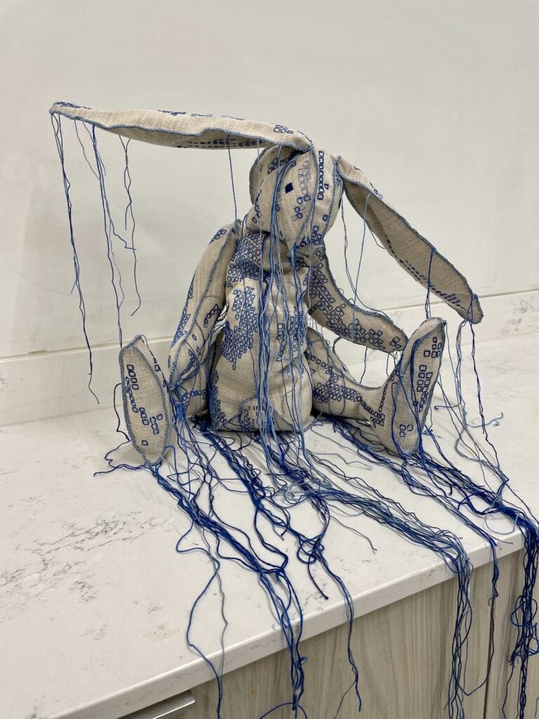 Sad looking rabbit 'stuffed animal' - embroidered with squares of different hues of blue, many strings hanging down from the fabric. Sitting on a white marble surface.