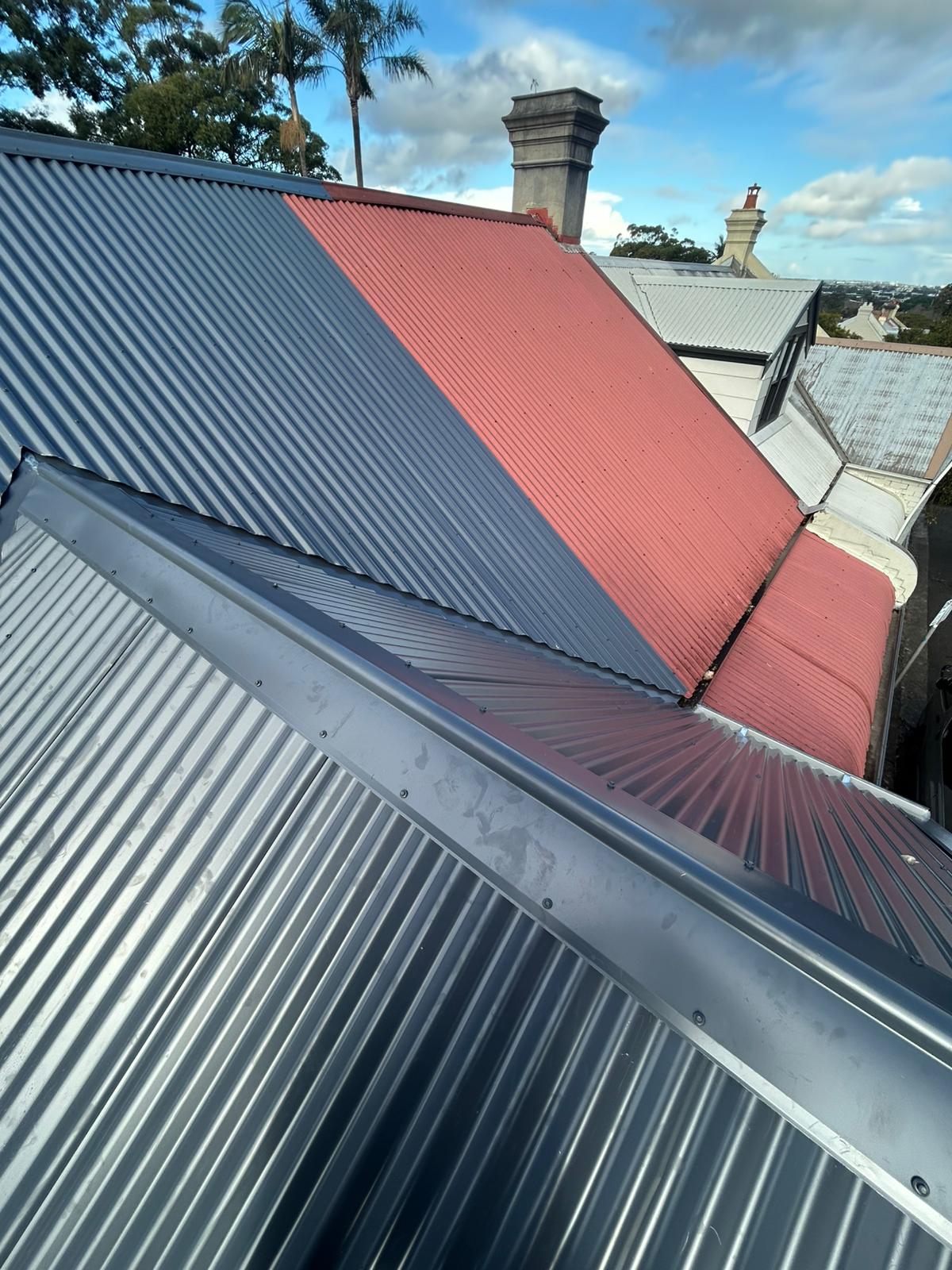 Corrugated metal roofing with ridge flashing, red and charcoal panels, chimneys in background.