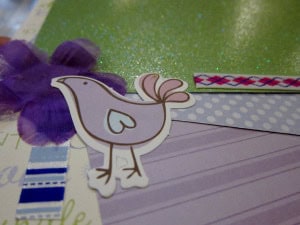 Baby scrapbook page with a purple bird sticker, purple flower, and various colored and patterned paper layers.