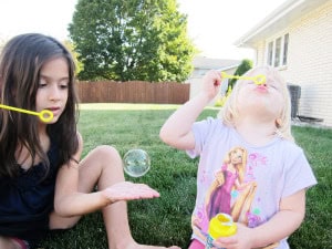 Two children sitting on grass blowing bubbles, one catching a bubble on her hand while the other blows through a wand.