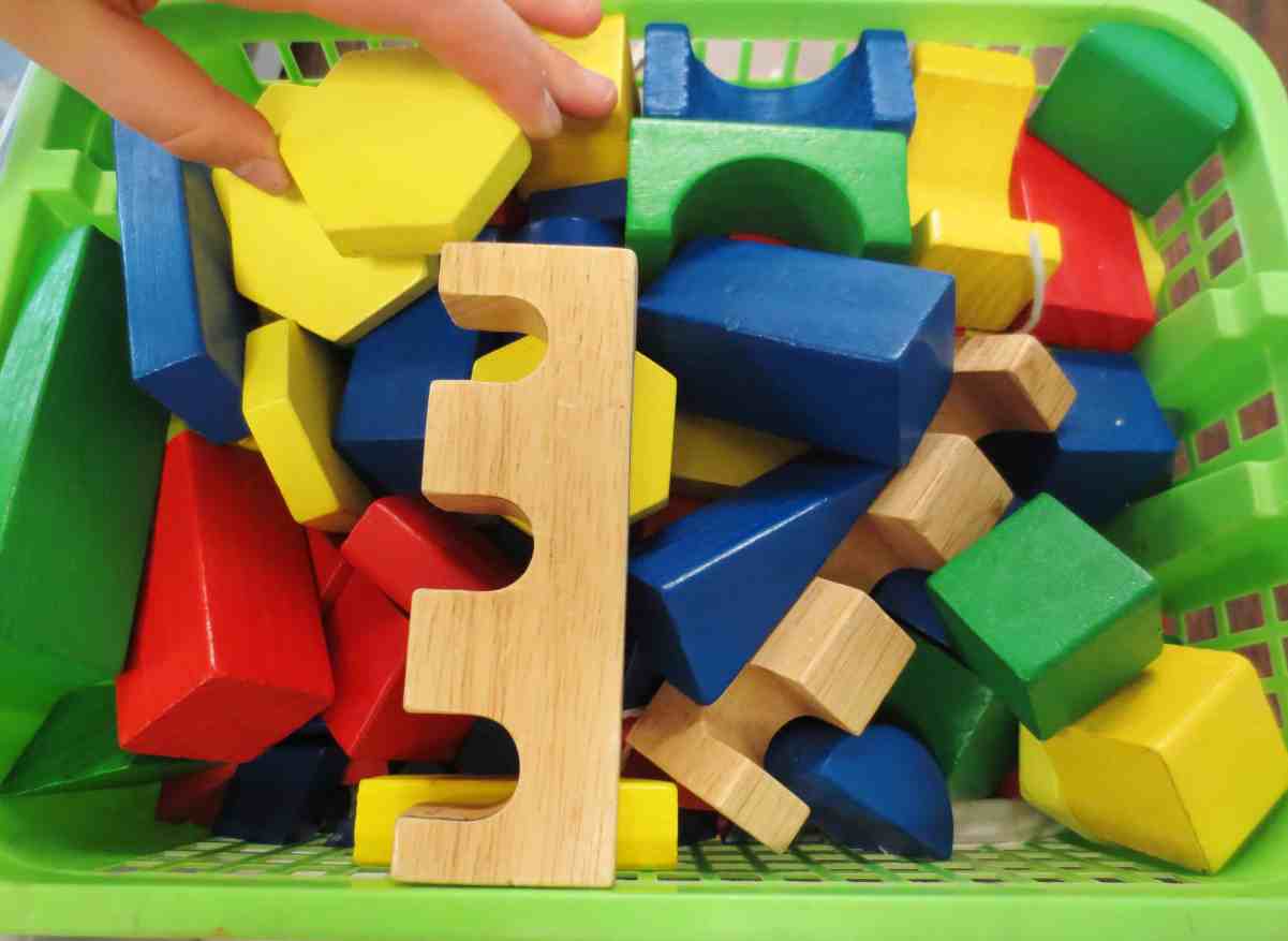 A colorful assortment of wooden pattern blocks in different shapes inside a green basket, with a hand reaching in.