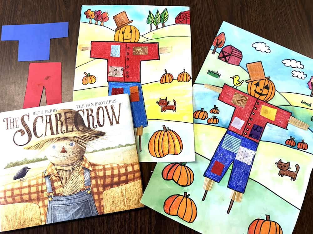 A book titled "The Scarecrow" sits beside two colored scarecrow drawings and a cut-out paper scarecrow template, perfect for a kids' scarecrow art or fall art lesson on a wooden table.