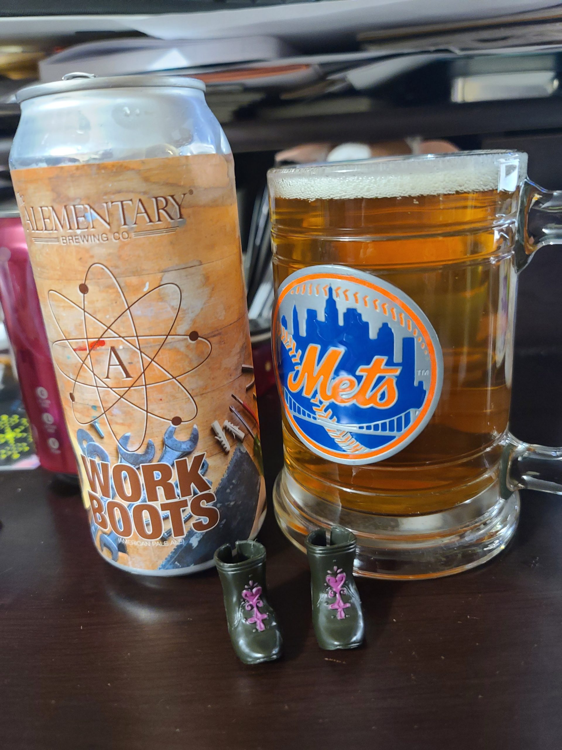 Mets stein filled with cream colored beer, with a small head, next to a tan can of Alementary Work boots and two doll shoes in front of it