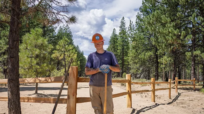 A young man stands proudly next to the new cedar fence he has built.