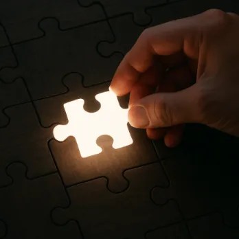 A single hand placing a glowing, white puzzle piece into a dark wooden jigsaw puzzle.