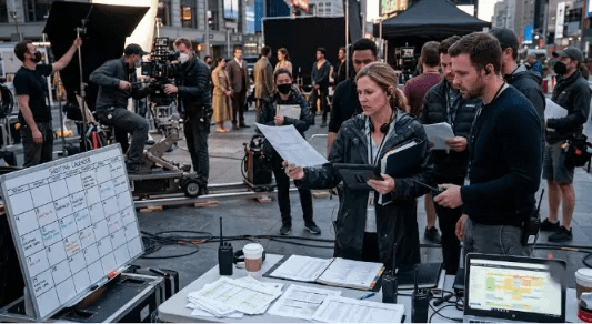 A film production crew working on a busy outdoor set with a whiteboard shooting calendar, paper schedules, and a laptop displaying project management software in the foreground.