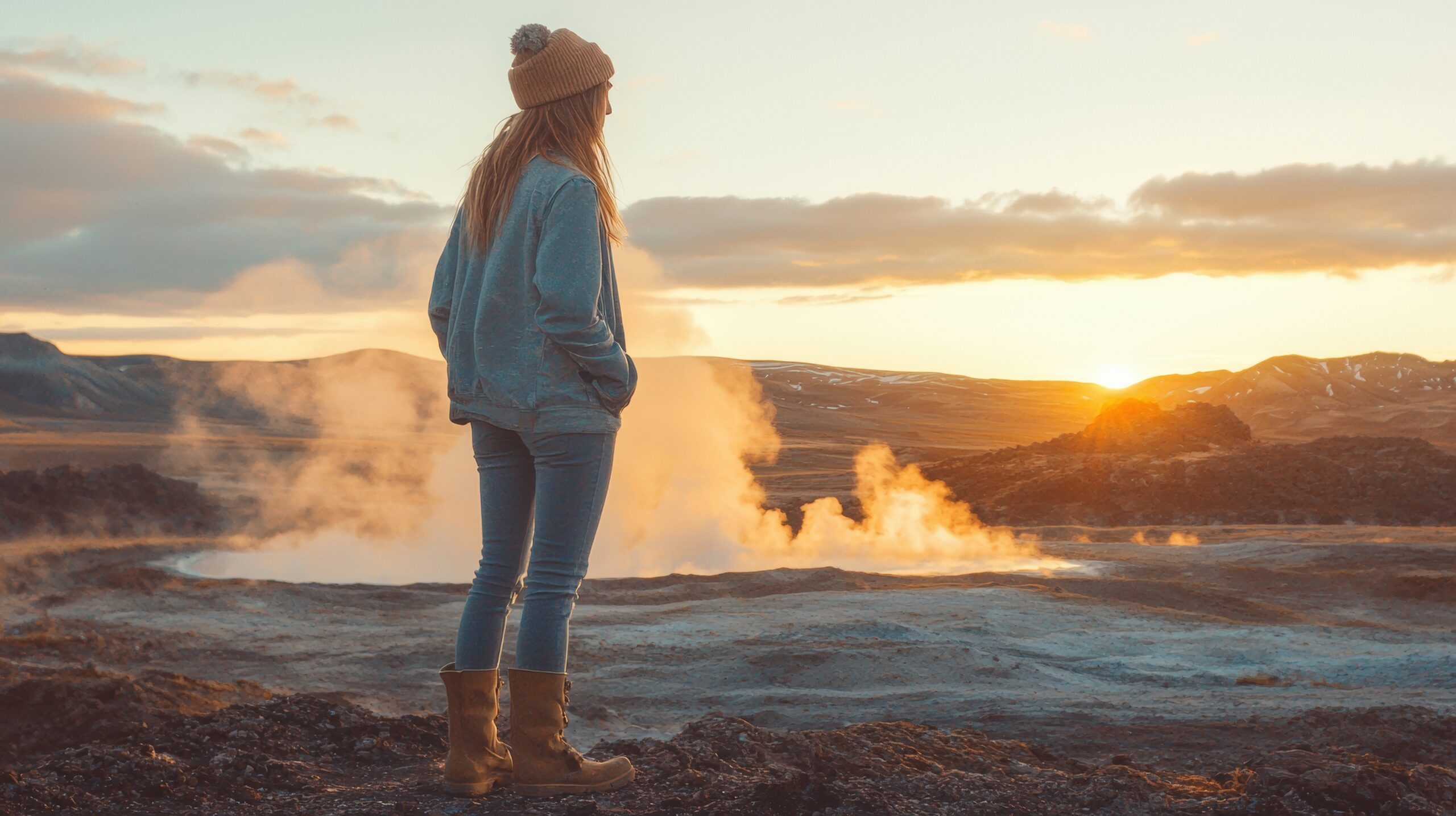 A woman hiker stands on a rocky ridge, gazing toward the horizon as the sun sets, bathing the landscape in warm orange and gold light.