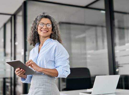 Smiling female CEO holding an iPad in a modern office setting, representing empowered empathetic leadership.
