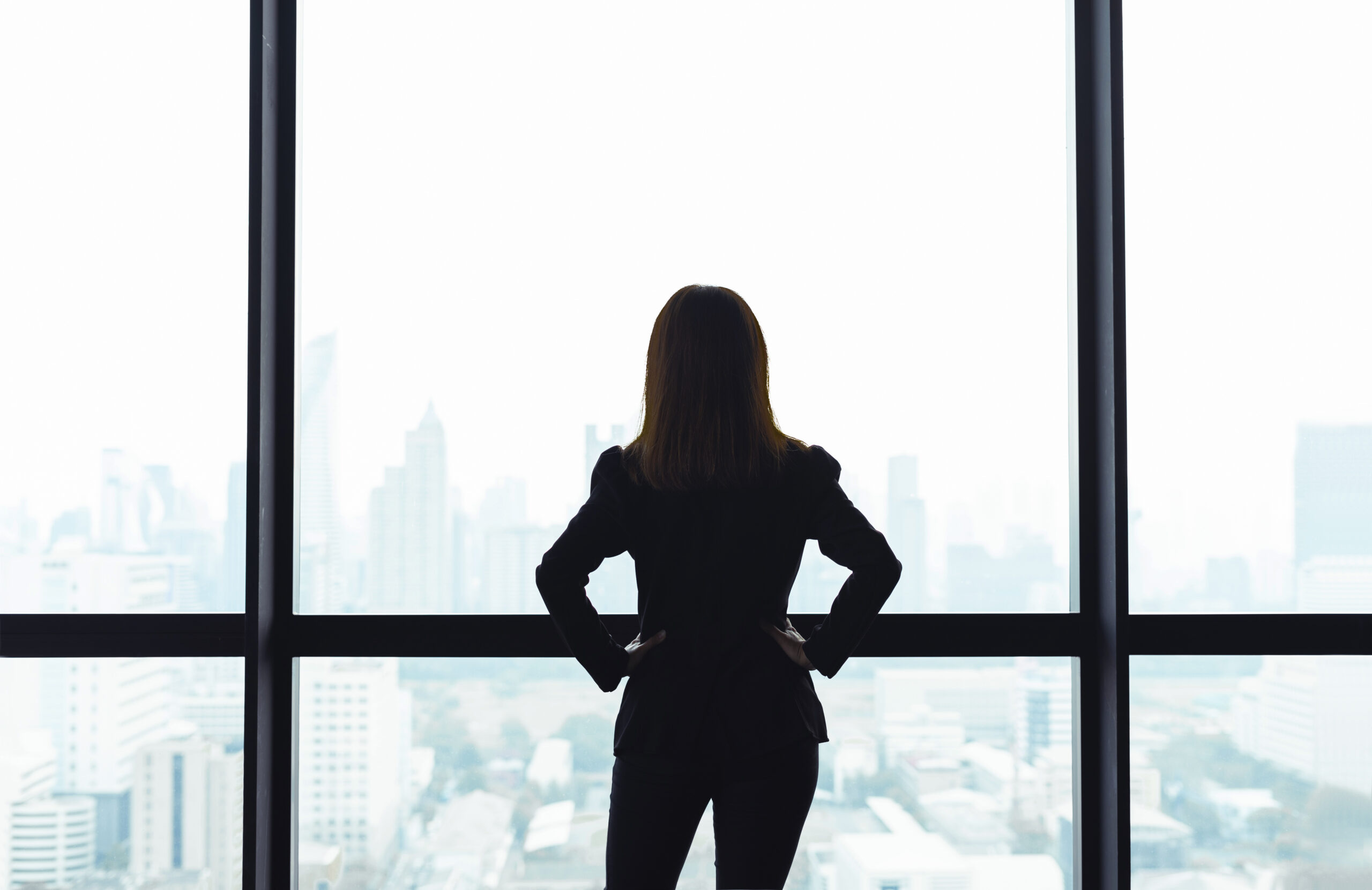 Confident female business leader standing at a high-rise window, overlooking a city skyline with hands on hips-symbolizing clarity, strength, and leadership.