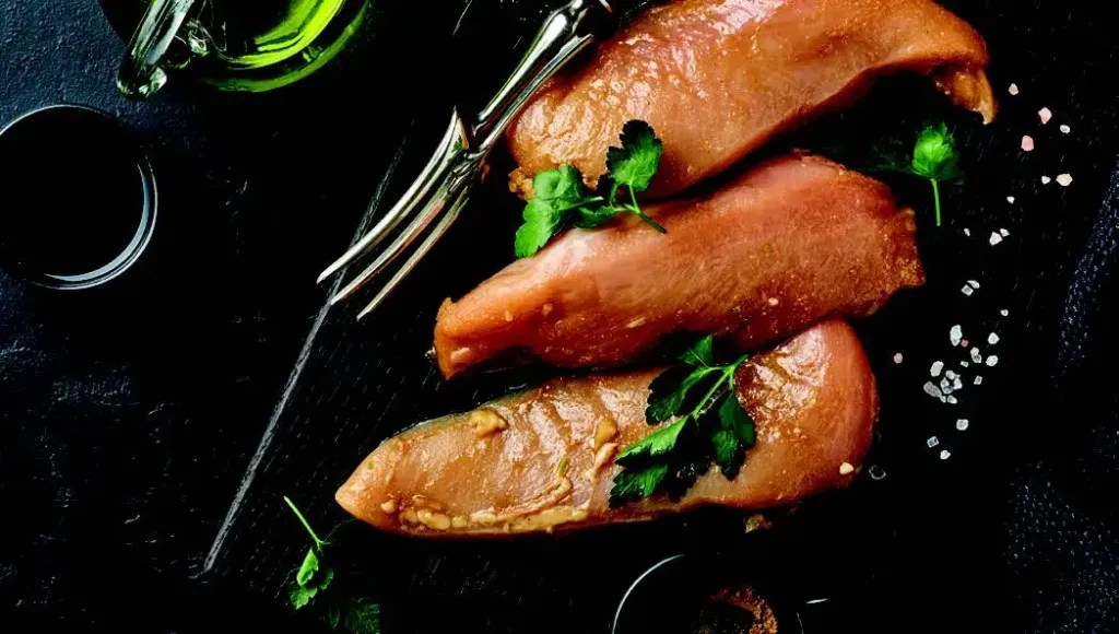 Raw marinated chicken breasts garnished with herbs on a dark surface