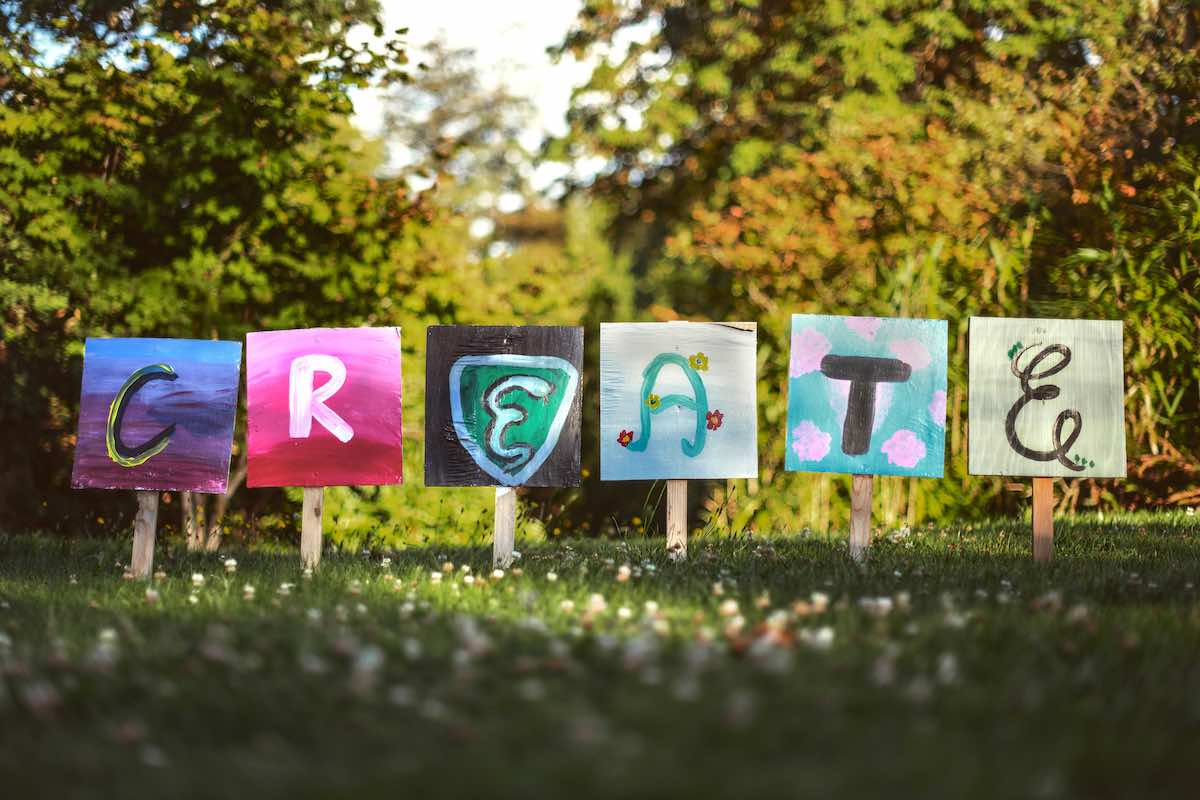 At the edge of a deciduous forest, six square boards on 2 by 4 stakes, spell out the word CREATE. Each board looks hand painted by kids. In the foreground a flower filled meadow.