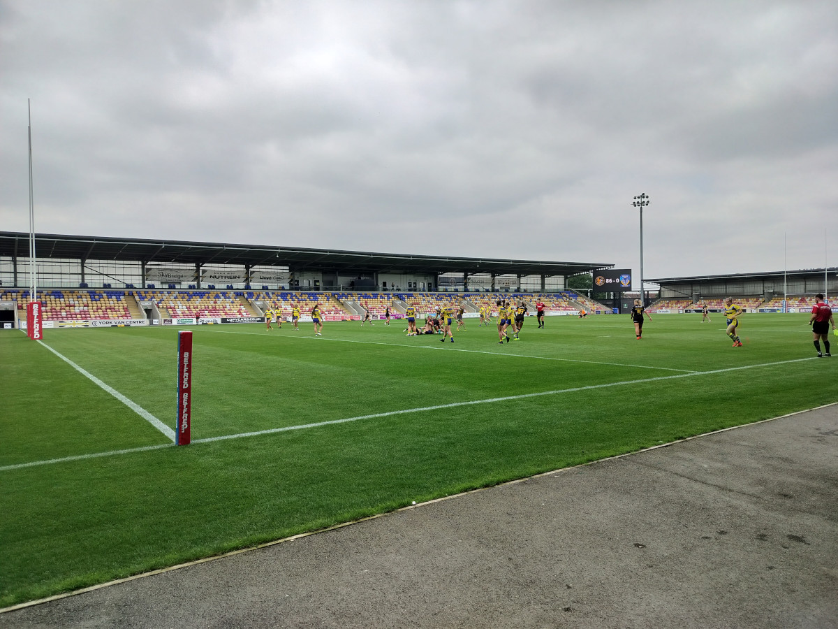View from the sidelines of a rugby league match taking place on the green grass of a small stadium on a cloudy day