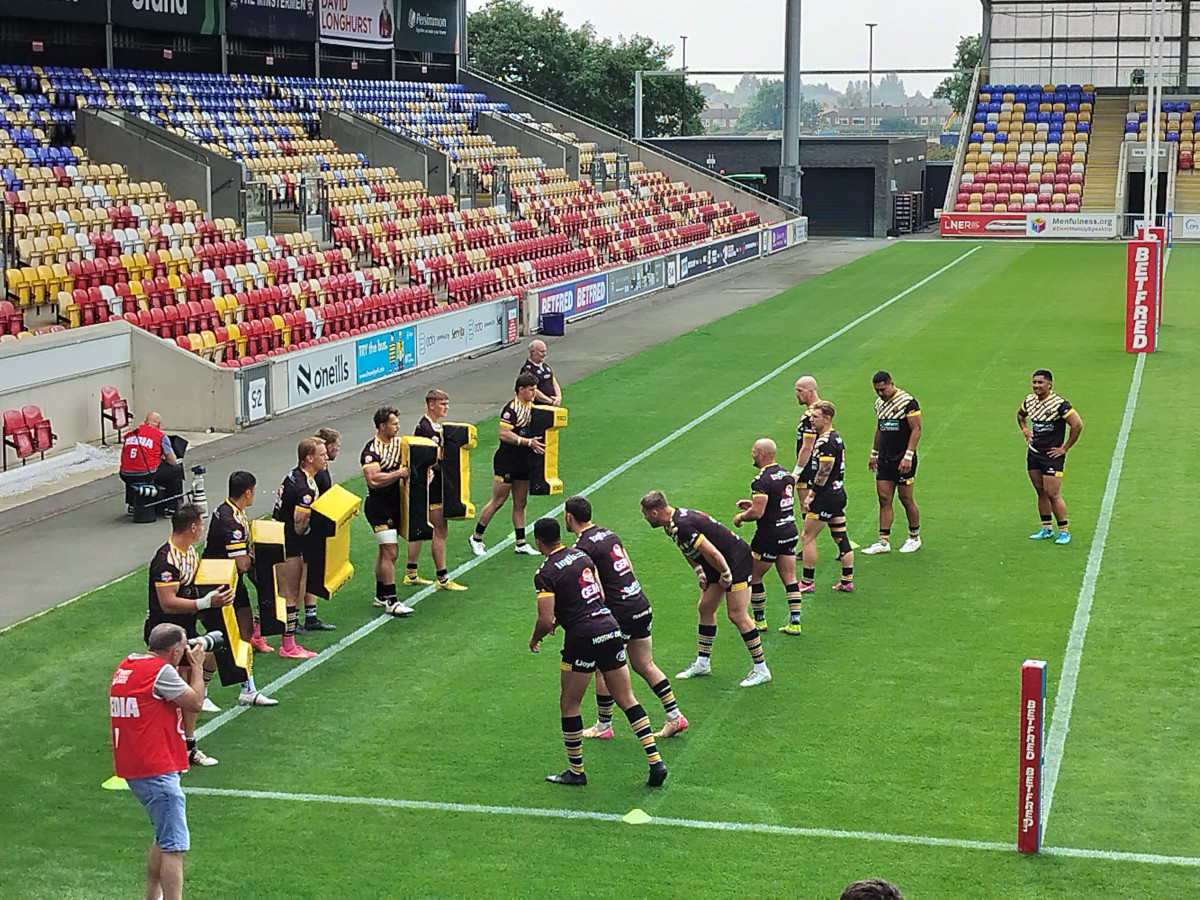 A men's rugby team warming up on the grass in a stadium. They are practicing tackles by running into large foam pads. Something you might see when Watching Rugby League Live For The First Time