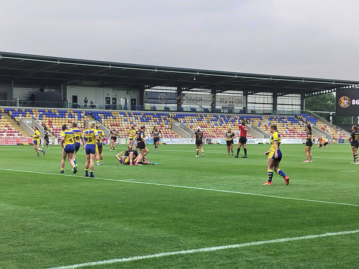 A ladies rugby league match in progress on a green pitch in a small stadium
