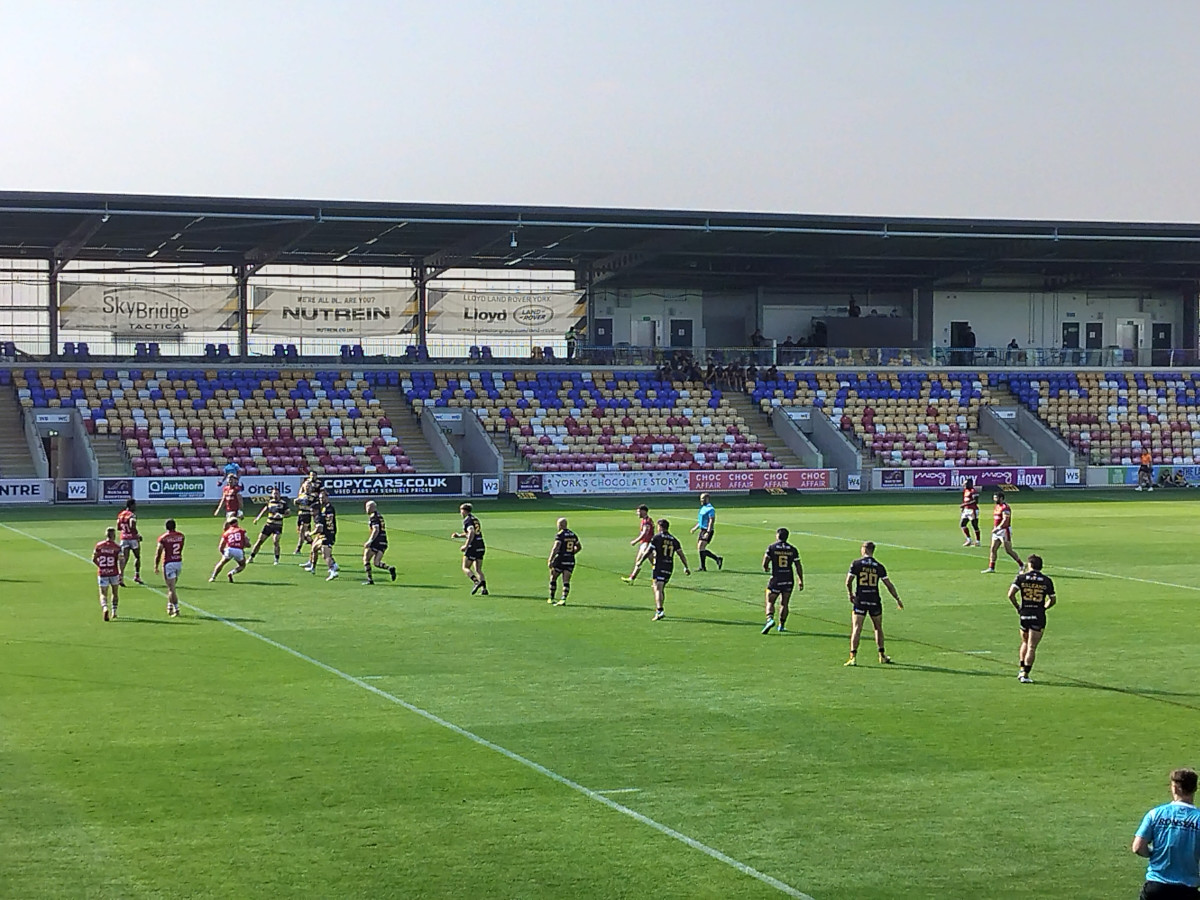 A men's rugby league match in progress on a green pitch at a small stadium.