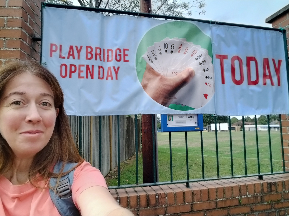 Challenge Fifty-Two, a white woman with dark hair smiling at the camera as she takes a selfie in front of a sign that reads 'play bridge open day today'