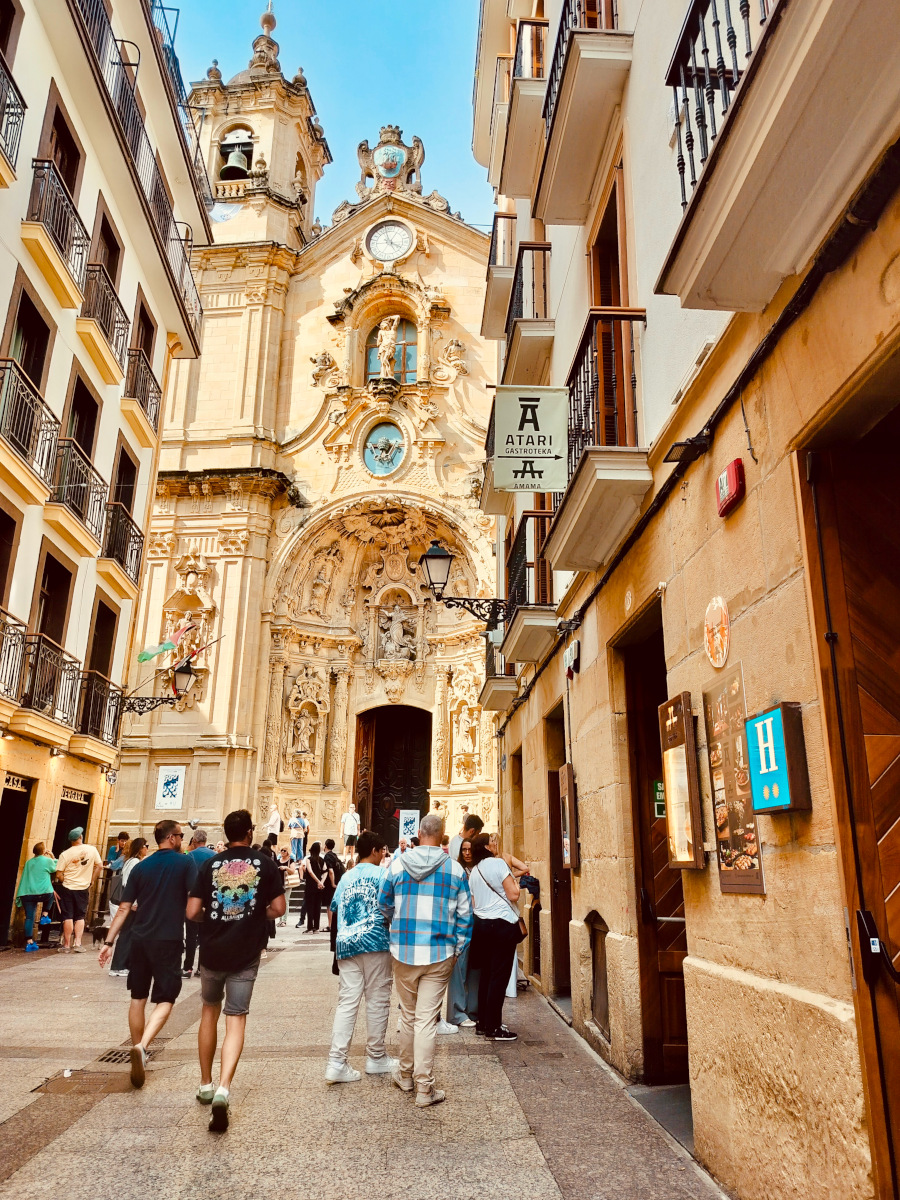 Looking along a street in San Sebastián on a Pintxos Tour. The Basilica of Santa María is visible at the end of the street. It's a nice day and there are a few people on the street.