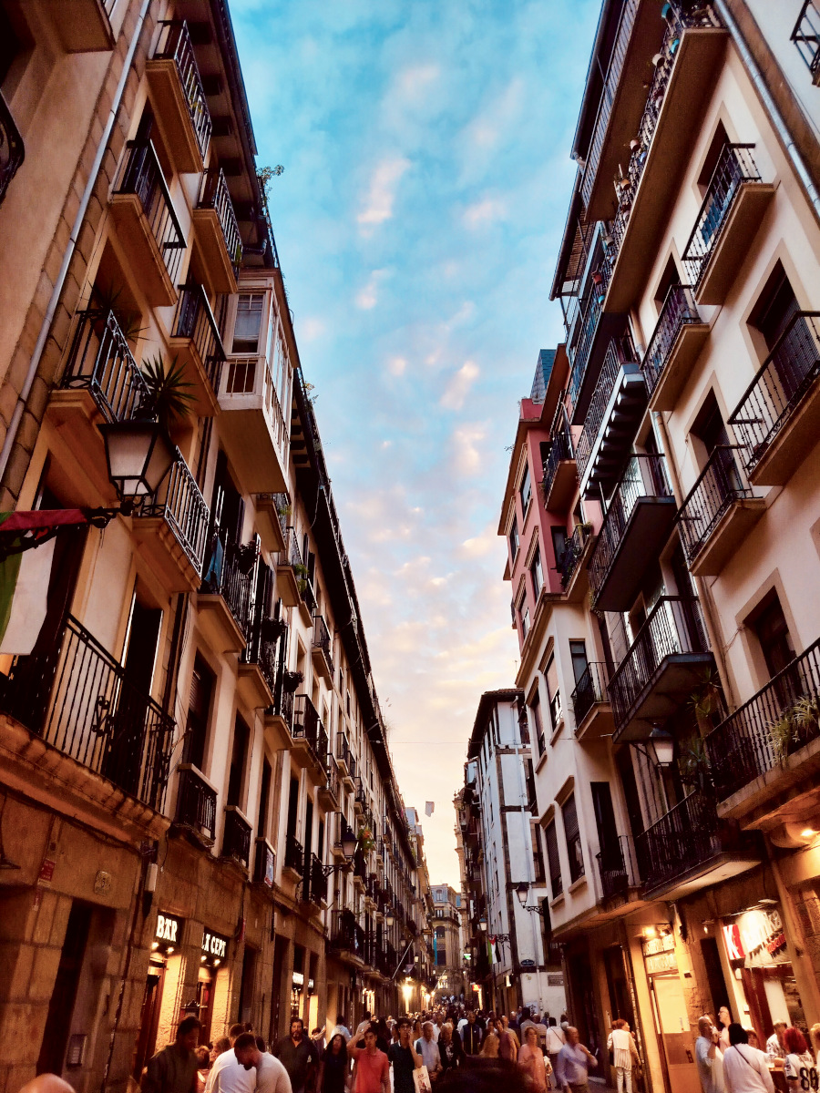 Looking along a narrow street in San Sebastián at evening. Lights are on in the bars and lots of people are milling about