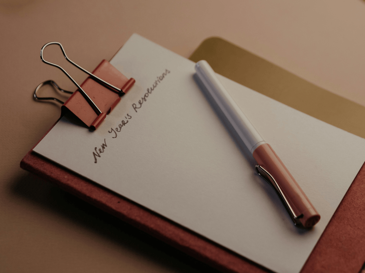 A note pad with neat handwriting at the top which reads 'new year resolutions' a pastel coloured pen lies across the paper