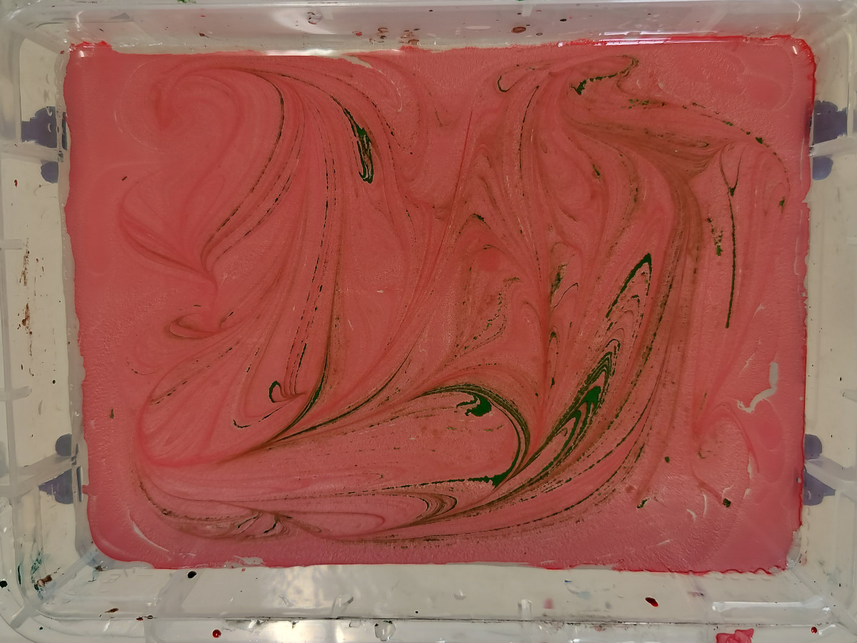 Looking down into a perspex container. There is liquid in it, but there is paint on the top. The paint has made a nice pink and green pattern. This is an example of trying bauble marbling