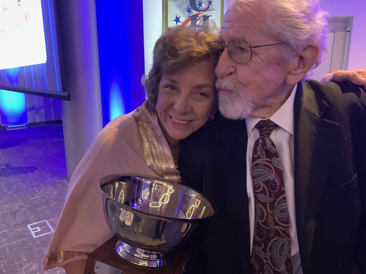 An older woman, Suzanne Pennink embraces her husband, Dr. Menno Pennink, while they pose for a photo with an award in their hands.