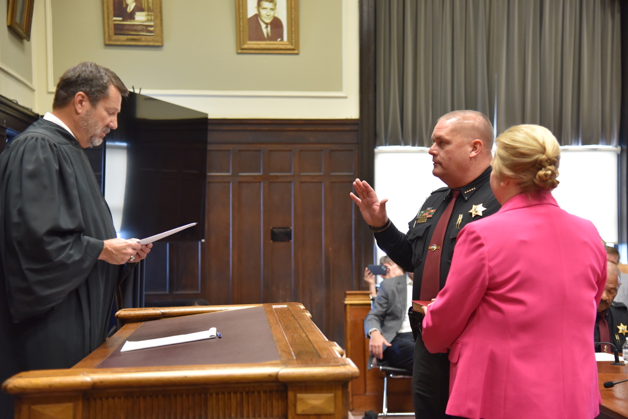 A white man in judge's robes is swearing in another white man wearing a police uniform and raising his right hand. The back of a woman wearing pink is visible.