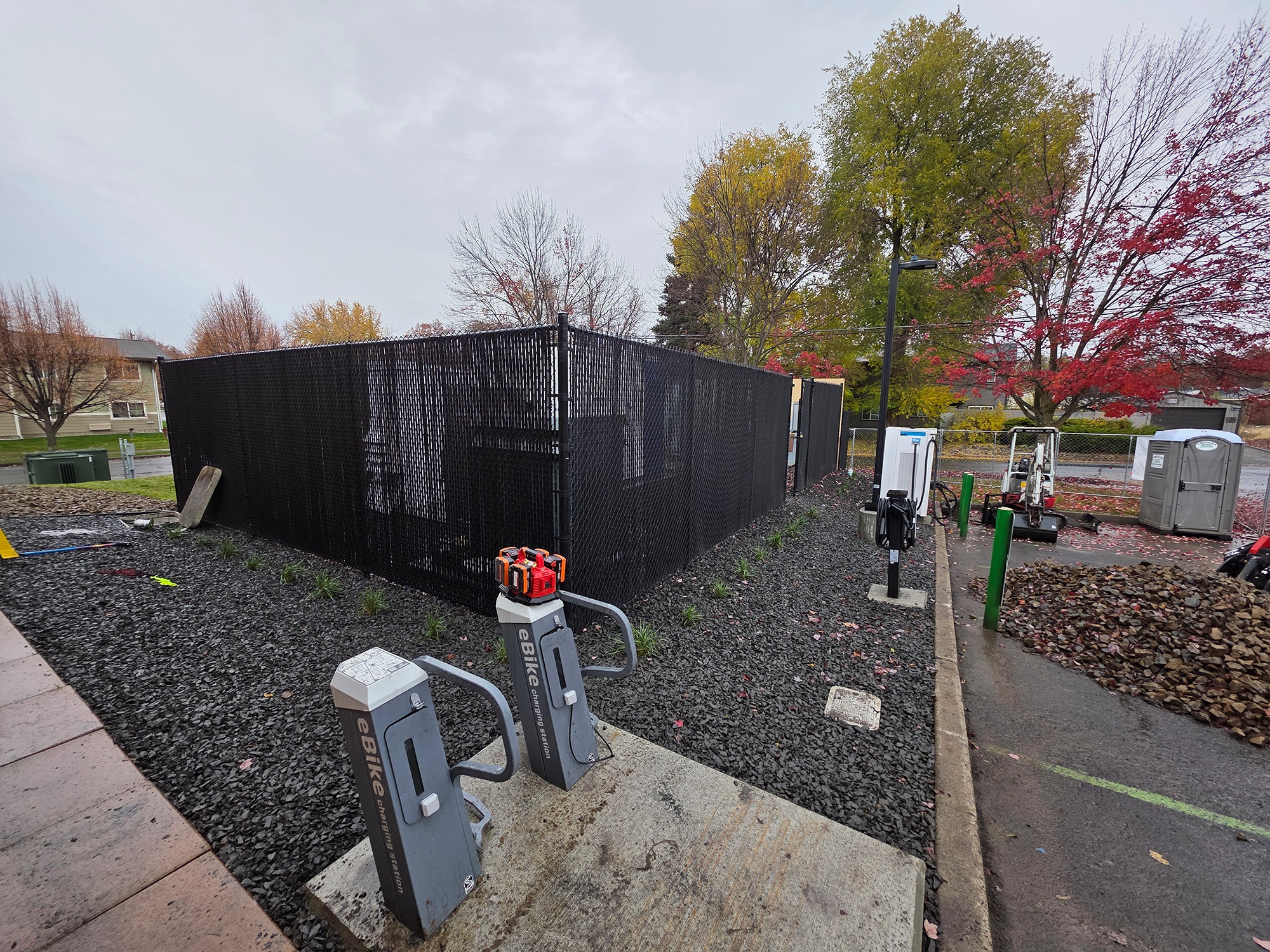 Fenced utility enclosure next to e-bike charging stations and parking area with autumn trees in the background.