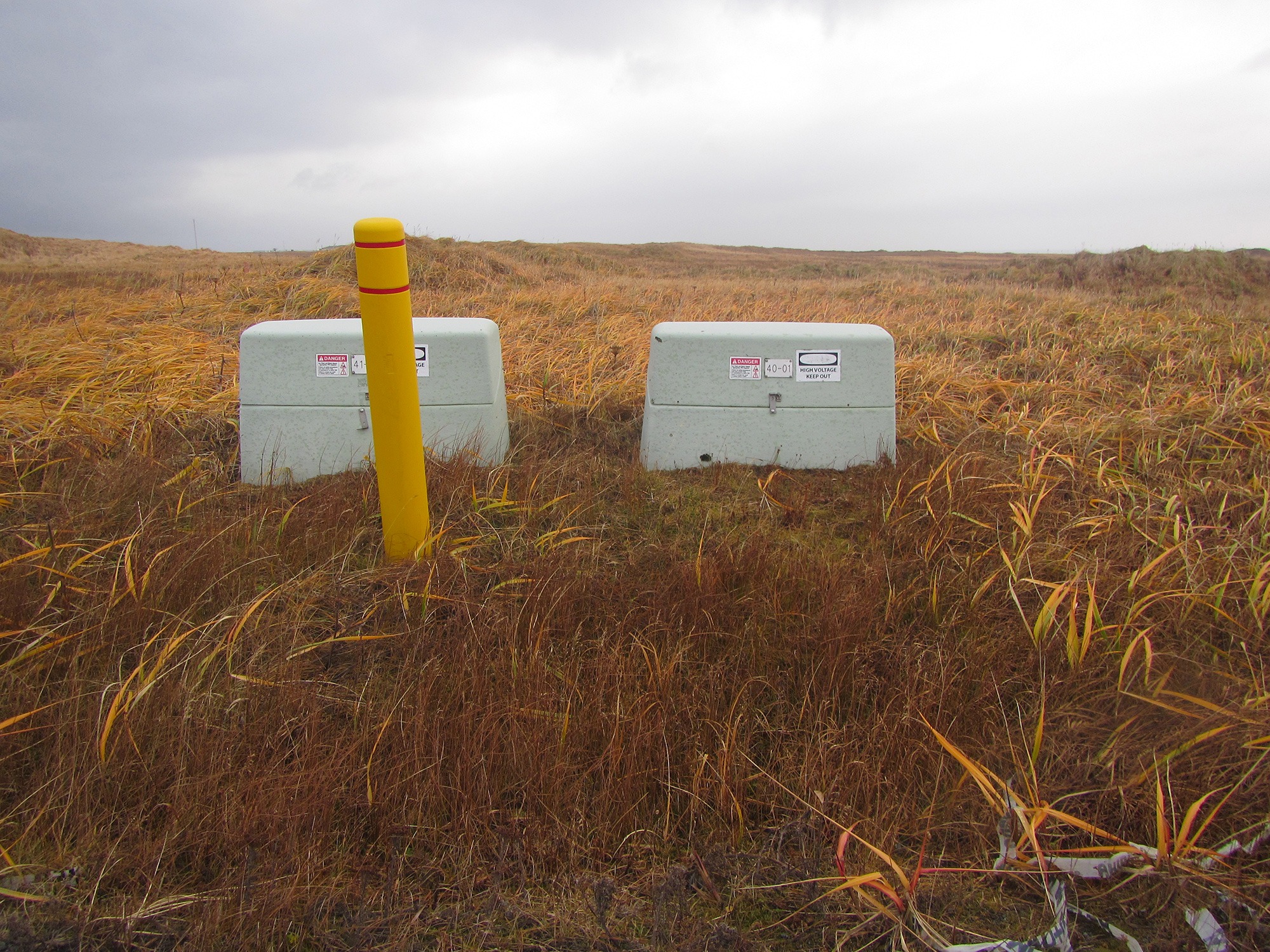 Two high-voltage electrical utility cabinets and a yellow protective bollard located in a grassy field under an overcast sky
