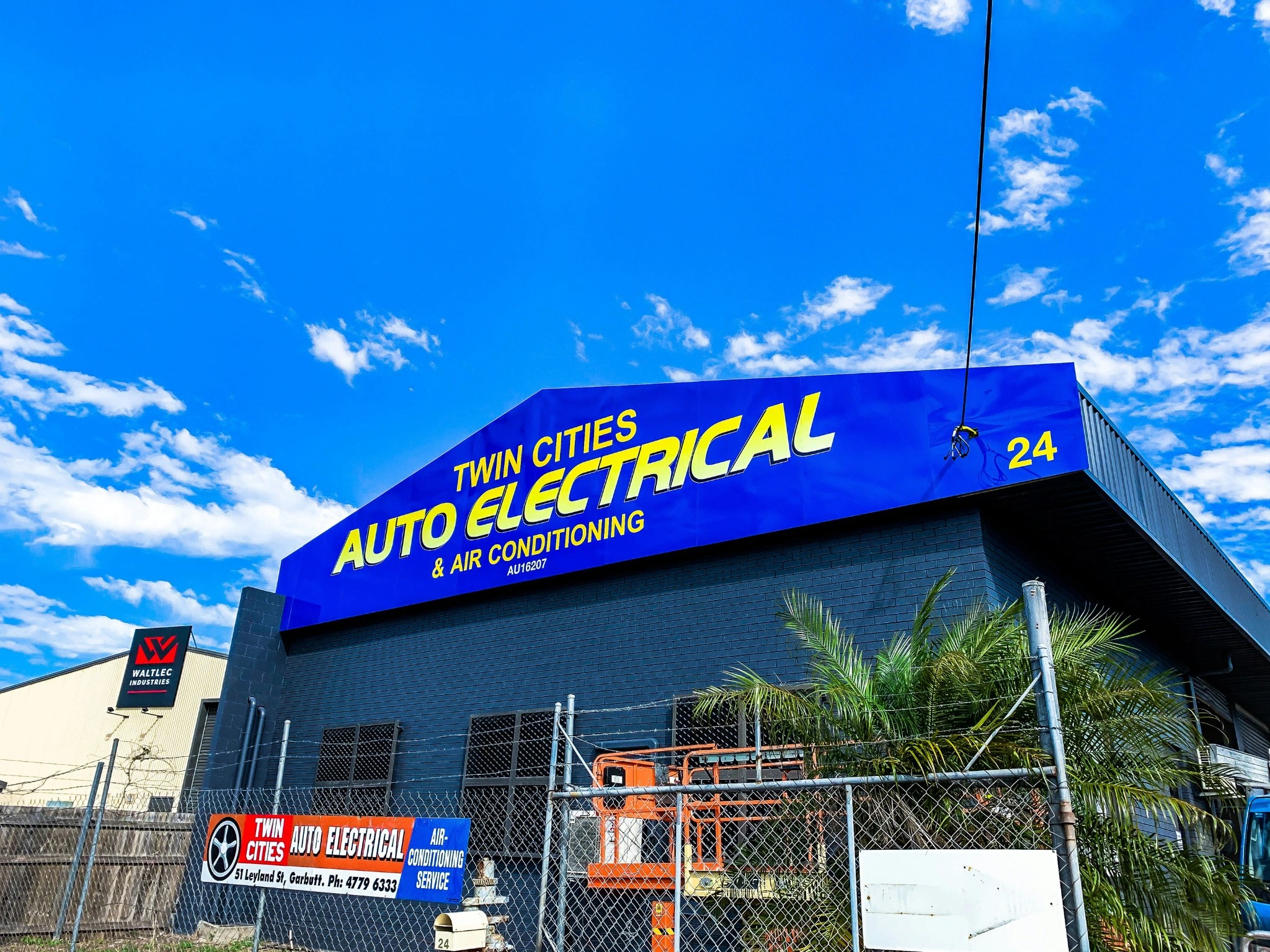 Commercial building with a bold blue sign reading 'Twin Cities Auto Electrical & Air Conditioning' under a bright blue sky