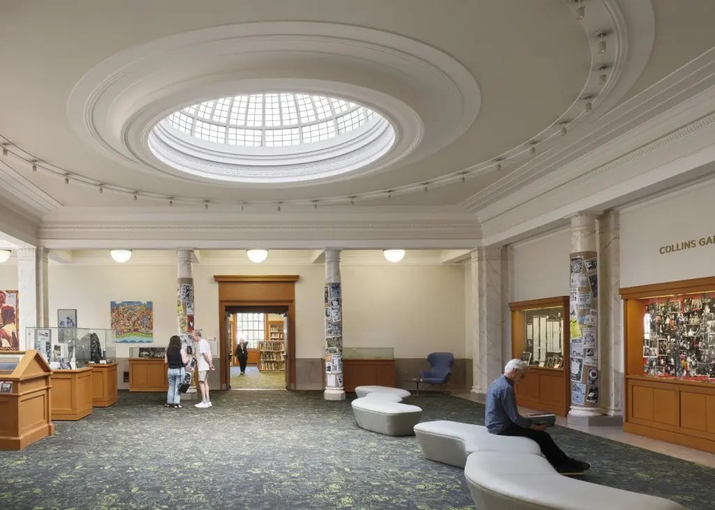 A library lobby with a domed skylight, columns, and art displays. Two people converse by a counter, while another sits on curved benches, creating a calm atmosphere.