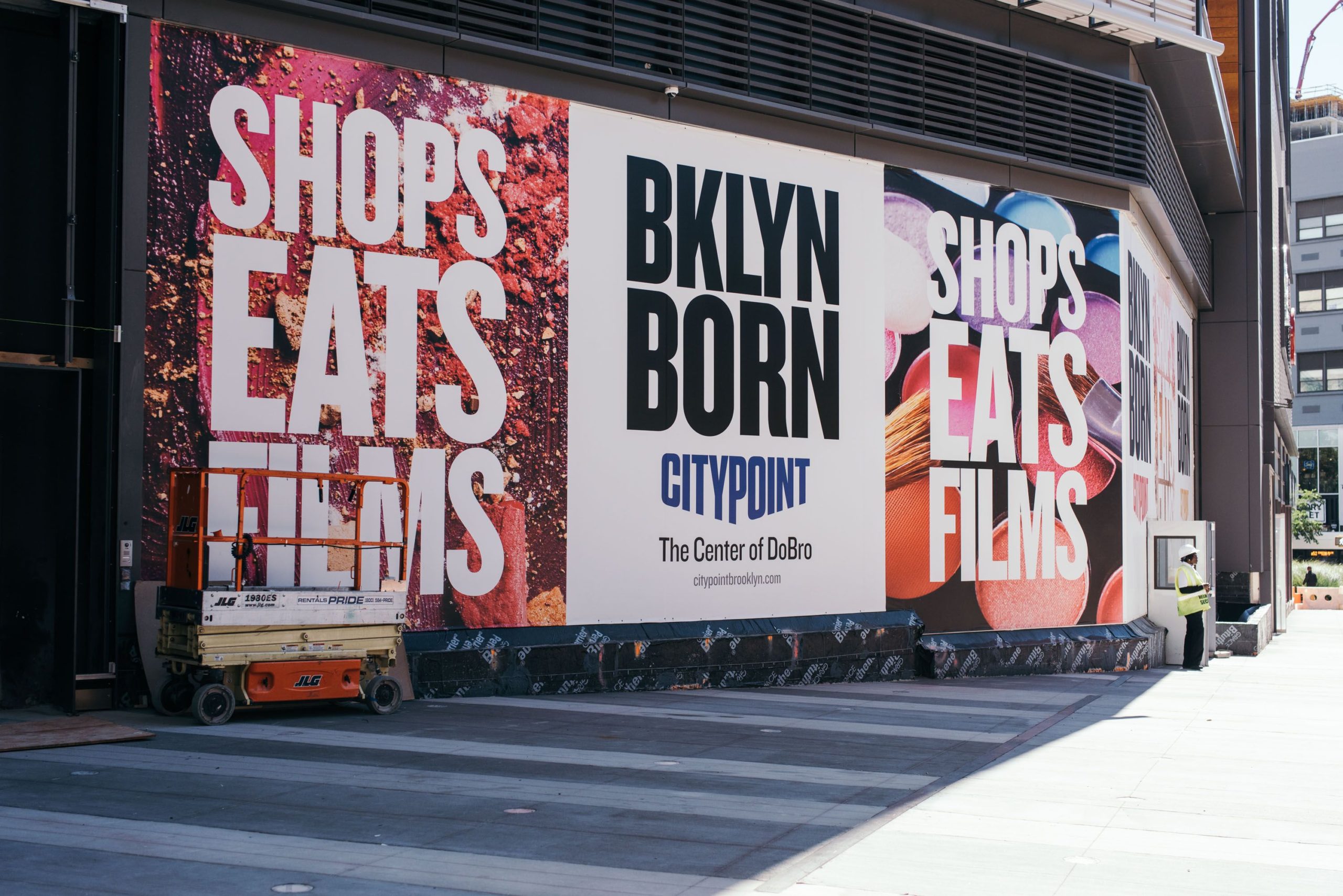 A vibrant outdoor wall wrap at CityPoint in Brooklyn, showcasing bold text promoting shops, eats, and films, alongside colorful images of various food items and products. A lift stands nearby, indicating ongoing installation or maintenance work.