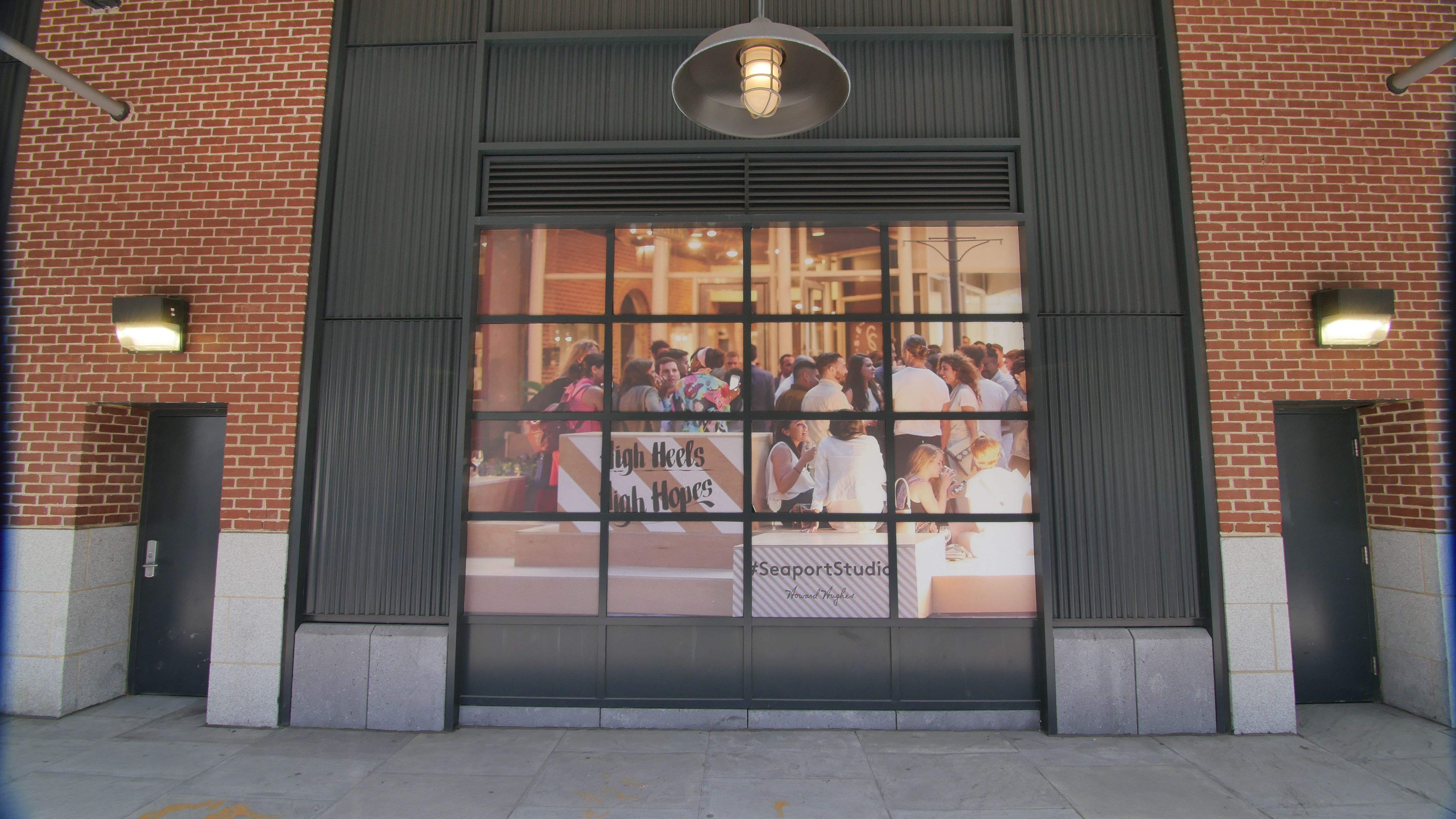 An exterior storefront featuring large glass windows displaying a busy scene of people engaged at an event or gathering inside, with vibrant decorations and signage indicating the theme 'Seaport Studios'. The brick wall and industrial-style elements of the building create a modern urban ambiance.