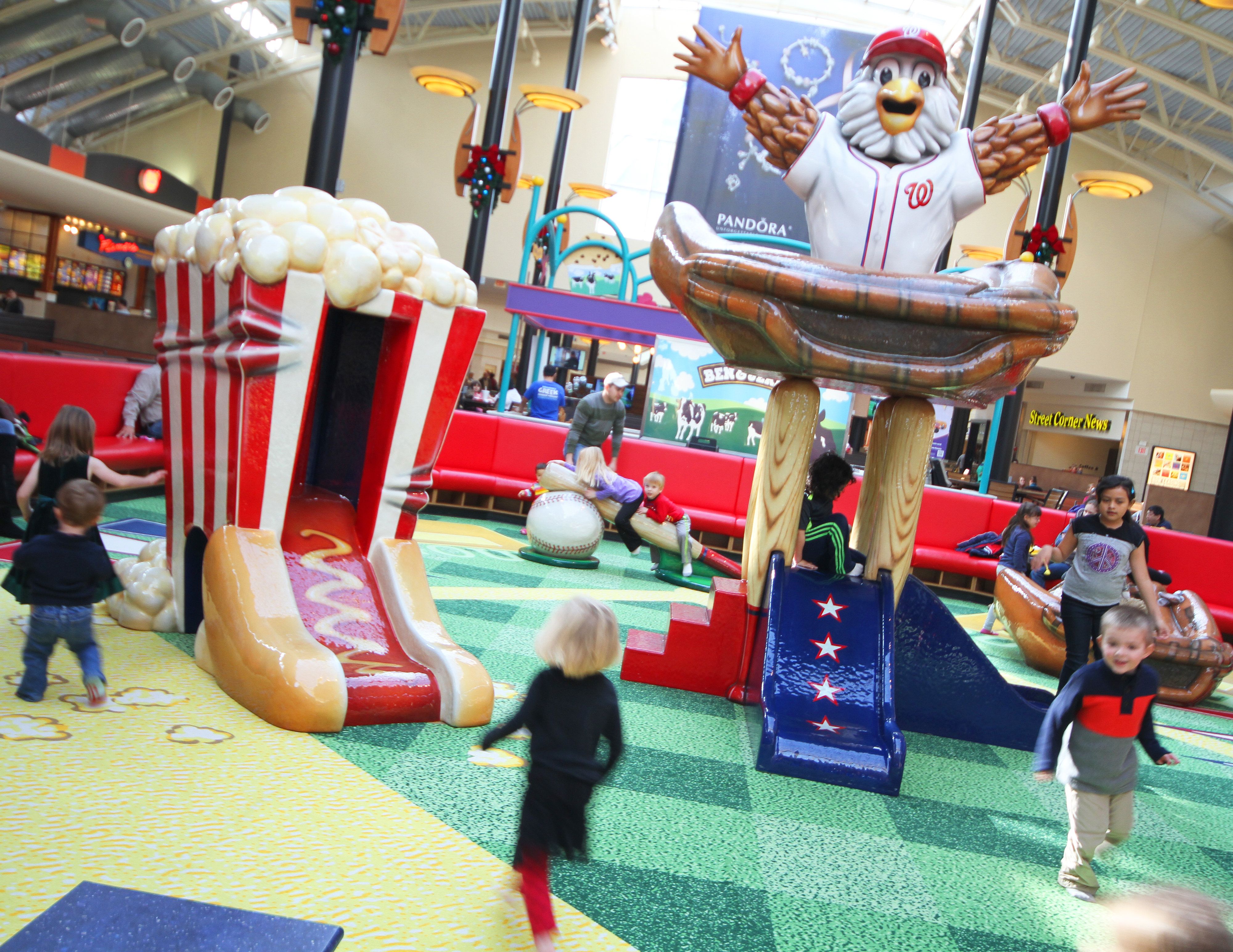 A vibrant indoor soft play park featuring a whimsical design with a giant popcorn container slide and a large chicken mascot in a baseball uniform. Children are playing and running around the colorful turf, engaging with the interactive elements. Bright decor and seating areas are visible in the background, creating a lively atmosphere.