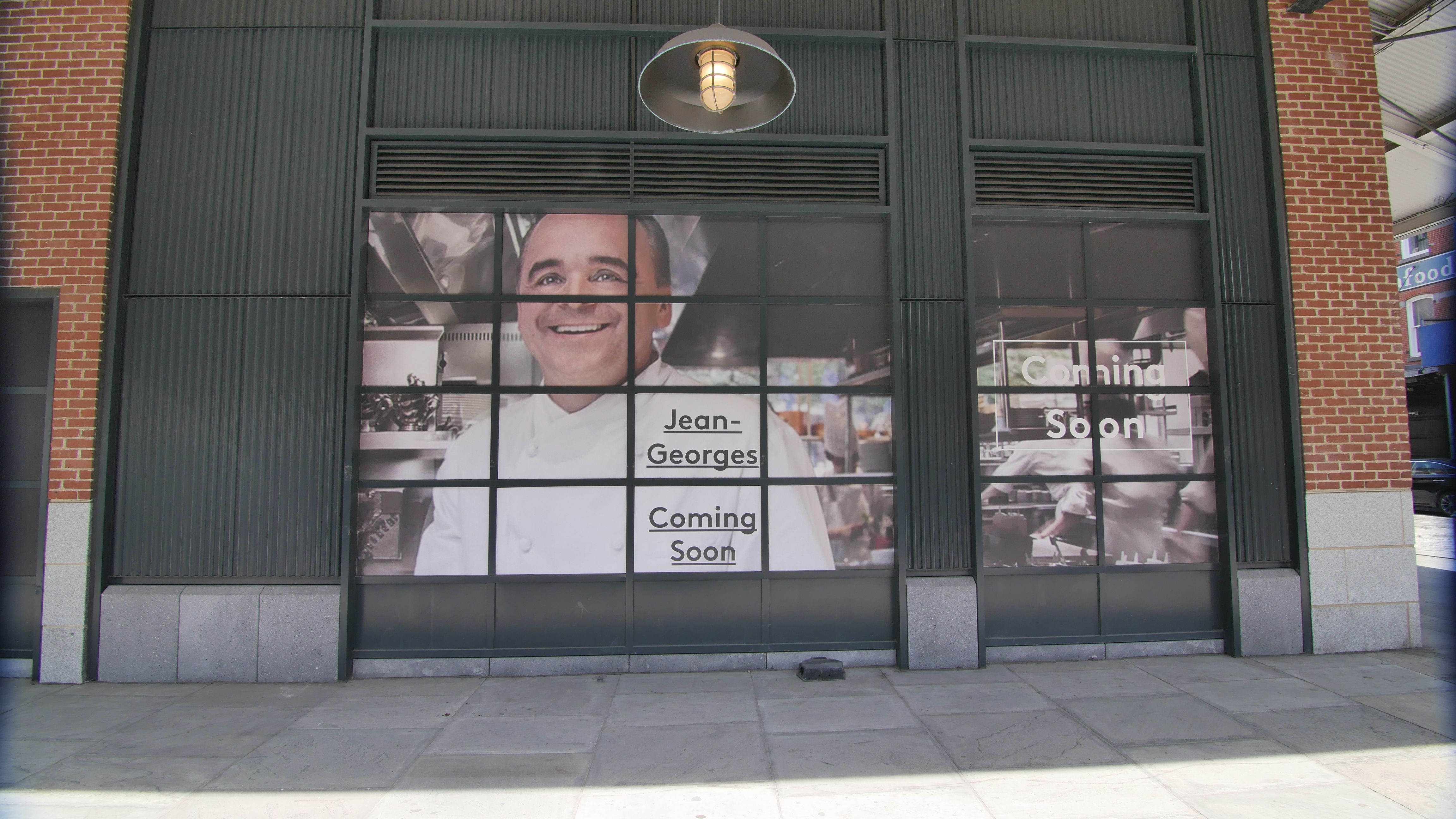 A large window display covered with promotional graphics for a new restaurant featuring a smiling chef's portrait and text that reads 'Jean-Georges Coming Soon.' The display's design uses bold typography against a blurred background of kitchen imagery, indicating an upscale dining experience.