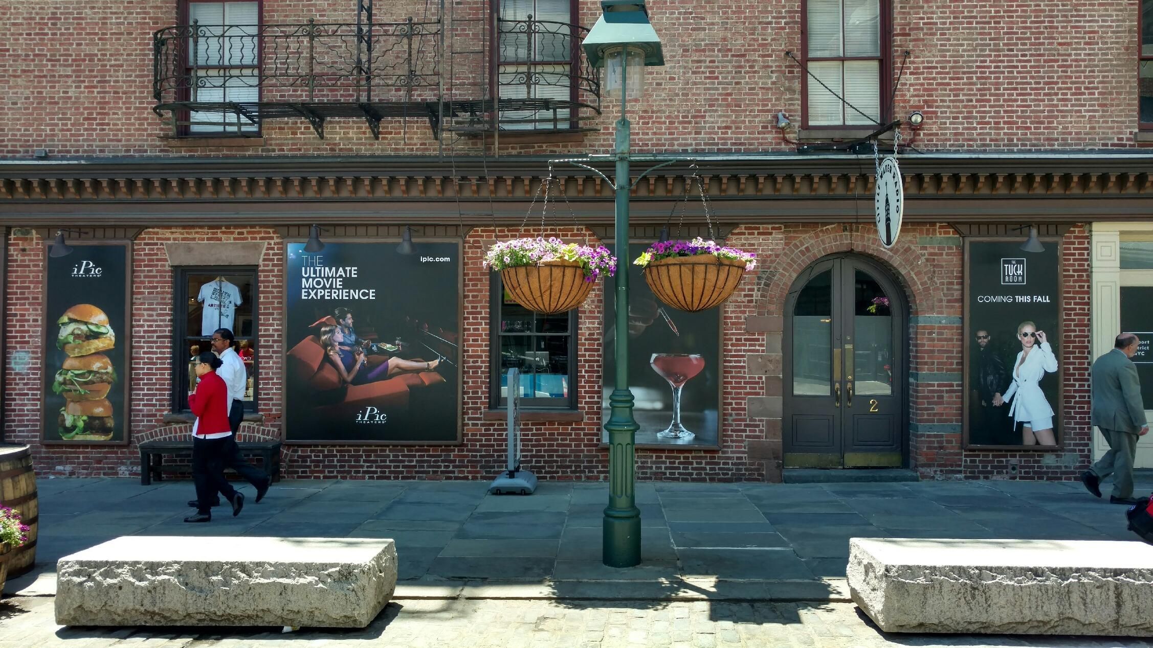 A bustling urban street scene featuring a historic brick building with large promotional posters for a movie experience, depicting food, drinks, and stylish individuals. Pedestrians walk by, and there are hanging flower baskets and benches in the foreground.