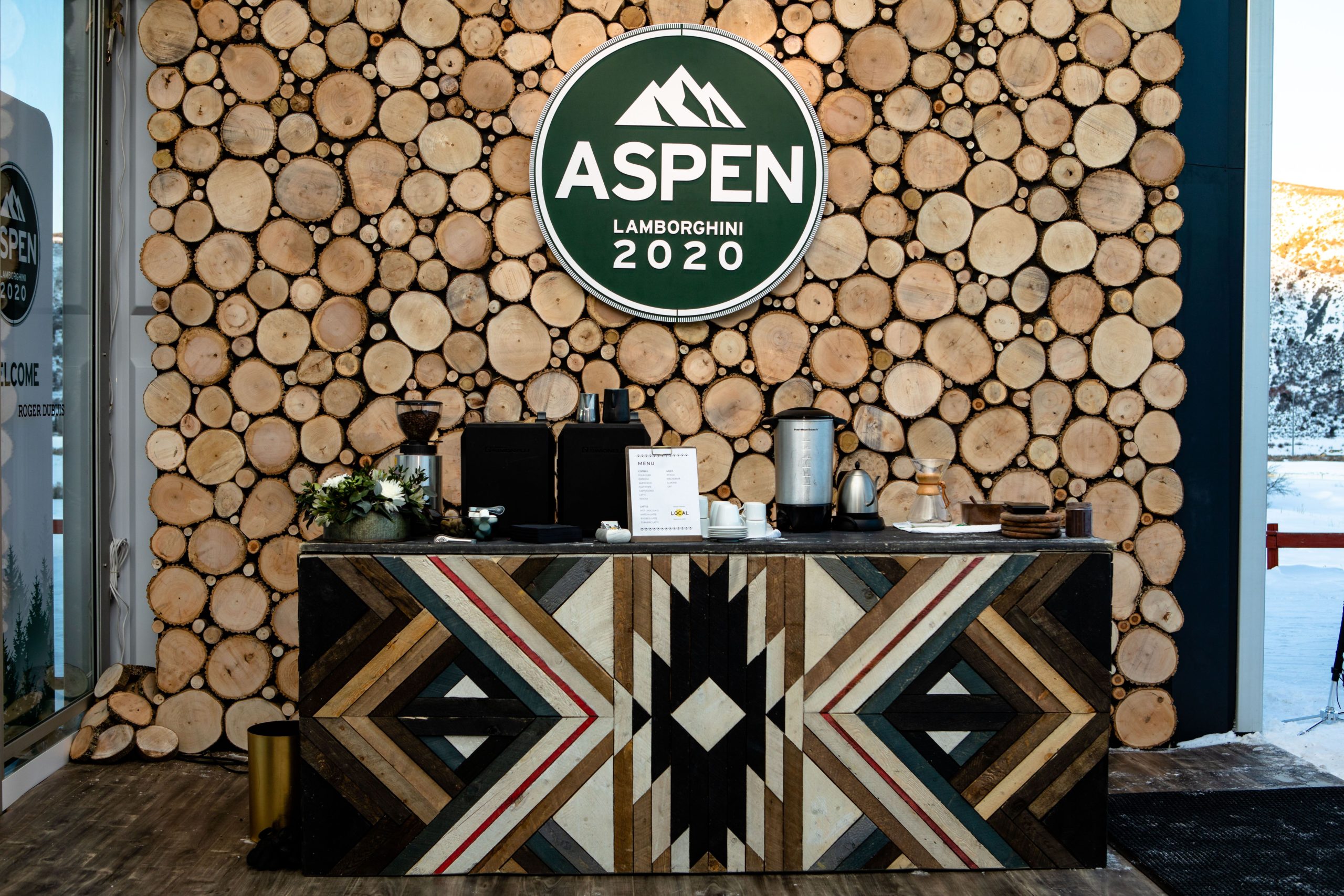 A stylish reception area featuring a wooden counter with an intricate geometric design, set against a backdrop of stacked logs. A circular sign above reads 'ASPEN LAMBORGHINI 2020'. Coffee equipment and a menu are placed on the counter, adding to the inviting atmosphere.