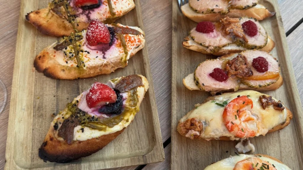 A wooden serving board displaying six gourmet tapas on toasted bread slices, featuring various toppings including sliced meat with strawberries and blackberries, prawns with melted cheese, and other colourful Mediterranean ingredients