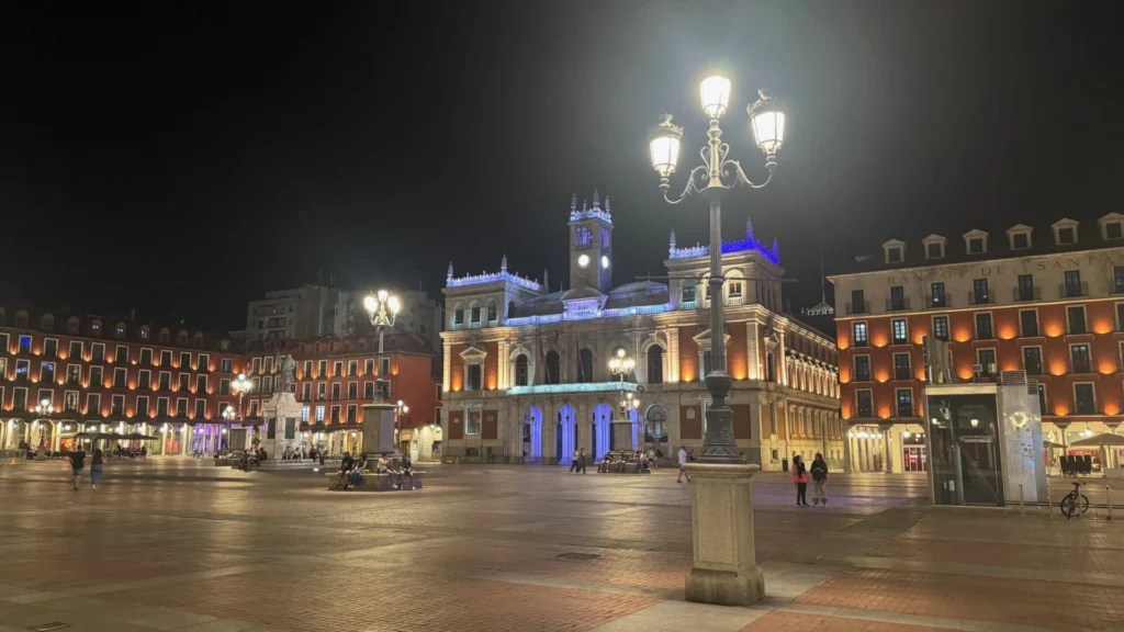 Night view of Plaza Mayor in Valladolid, Spain, showing the illuminated town hall with blue lighting and clock tower, surrounded by traditional red-brick buildings with warm golden lighting, ornate street lamps, and people strolling across the spacious cobblestone square