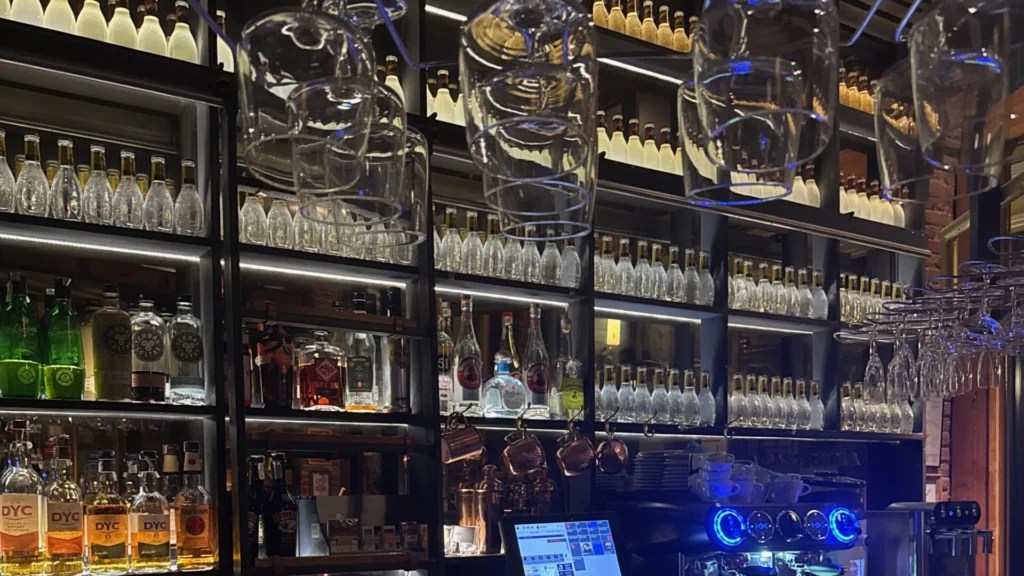 Interior view of La Sastrería cocktail bar showing a well-stocked bar with illuminated shelving displaying premium spirits and bottles, hanging wine glasses, a modern point-of-sale system, and professional bar equipment with blue LED lighting